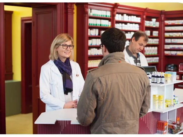 Eine Apothekerin berät einen Kunden an der Tara, im Hintergrund ein Apotheker