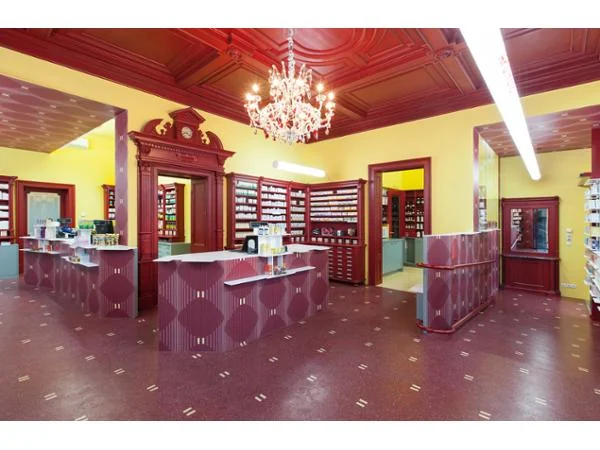 Verkaufsraum der Apotheke mit roter Kassettendecke, Kristallluster, historischen Regalen und gelben Wänden